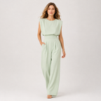 A woman modeling a light sage green sleeveless jumpsuit with a structured draped bodice and wide legs, standing against a plain light gray studio background.