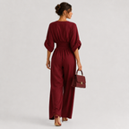 A woman is shown from behind wearing a deep burgundy jumpsuit with three-quarter sleeves and wide legs, holding a matching burgundy handbag against a light gray studio background.