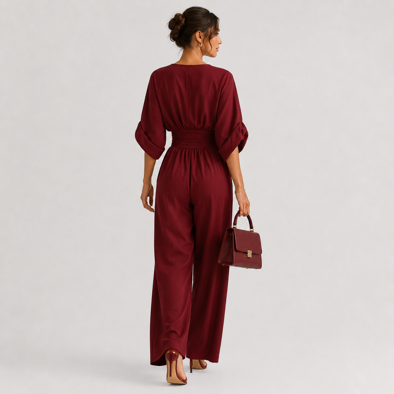 A woman is shown from behind wearing a deep burgundy jumpsuit with three-quarter sleeves and wide legs, holding a matching burgundy handbag against a light gray studio background.