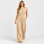 A woman modeling a sleeveless beige jumpsuit with a mock neck and wide legs, standing against a light gray studio background.
