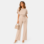 A woman models a beige short-sleeve jumpsuit with a tie waist, paired with a matching beige handbag and nude heels, against a plain white studio background.