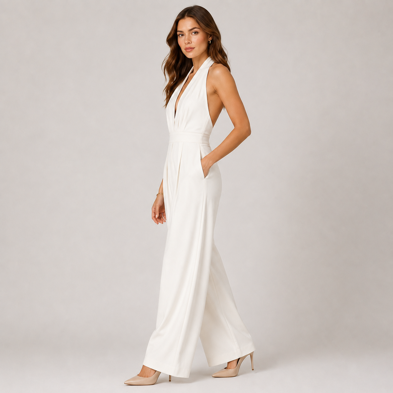 A woman modeling a white sleeveless jumpsuit with a deep V-neckline, side pockets, and wide-leg pants, paired with beige heels, against a light gray studio background.