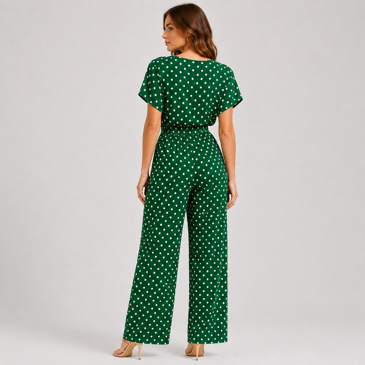 A woman modeling a green short-sleeve jumpsuit with a white polka dot pattern, shown from a rear three-quarter angle against a light gray studio background.