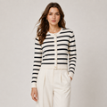 A woman modeling a black and white horizontally striped cardigan with gold buttons, paired with cream-colored trousers, against a neutral light gray background.