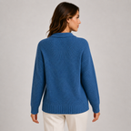 A woman shown from the back wearing a blue ribbed knit sweater and white pants, standing against a plain light gray studio background.