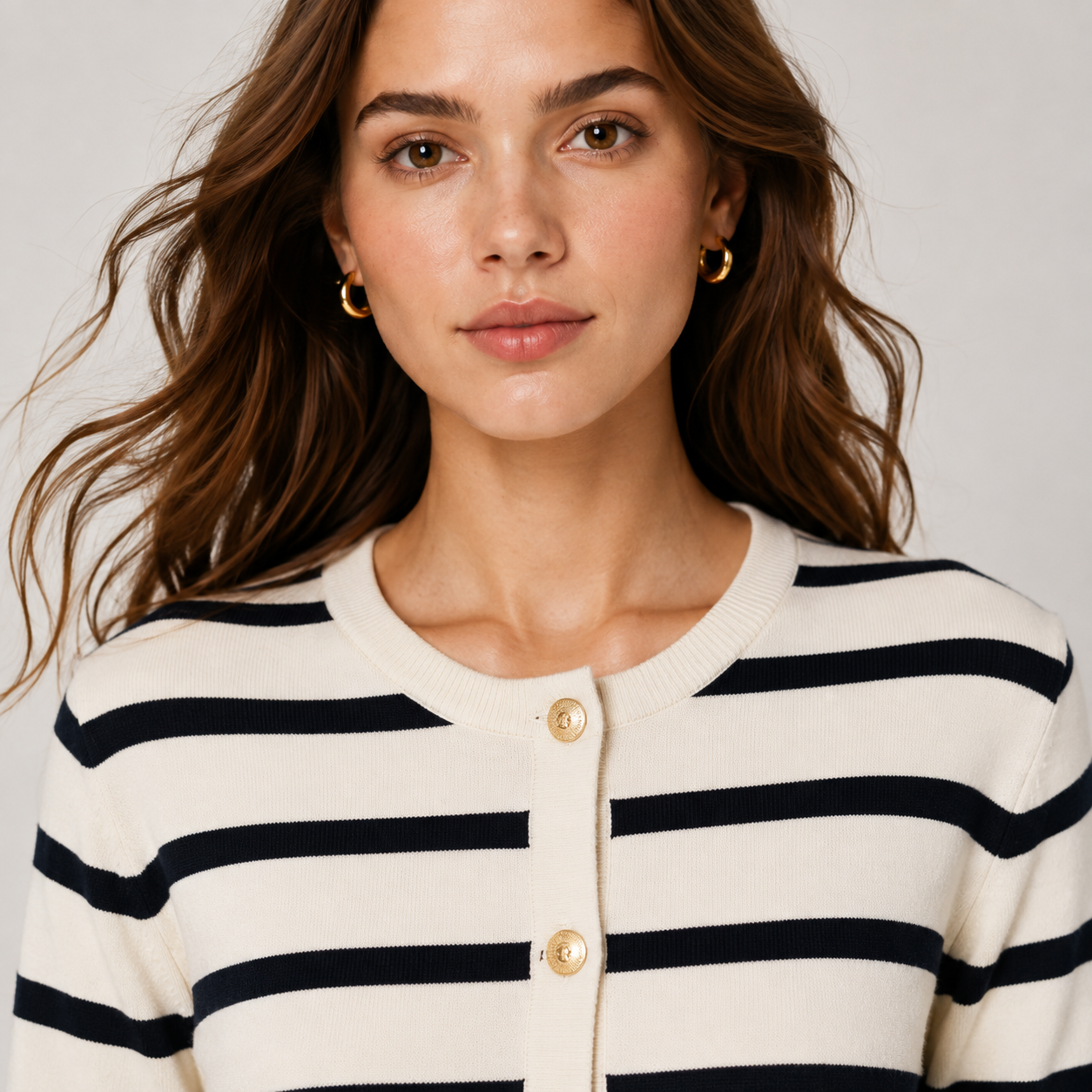 A close-up of a woman wearing a cream-colored knit cardigan with navy blue horizontal stripes and gold buttons. She is also wearing small gold hoop earrings, set against a plain light gray background.
