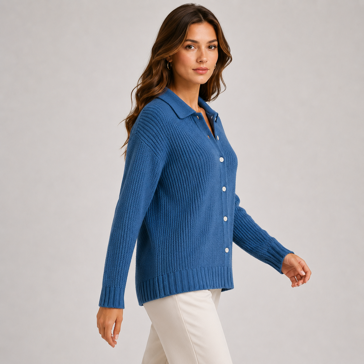 A woman wearing a blue ribbed knit button-front cardigan with a collar, paired with white pants, photographed against a plain light gray studio background.