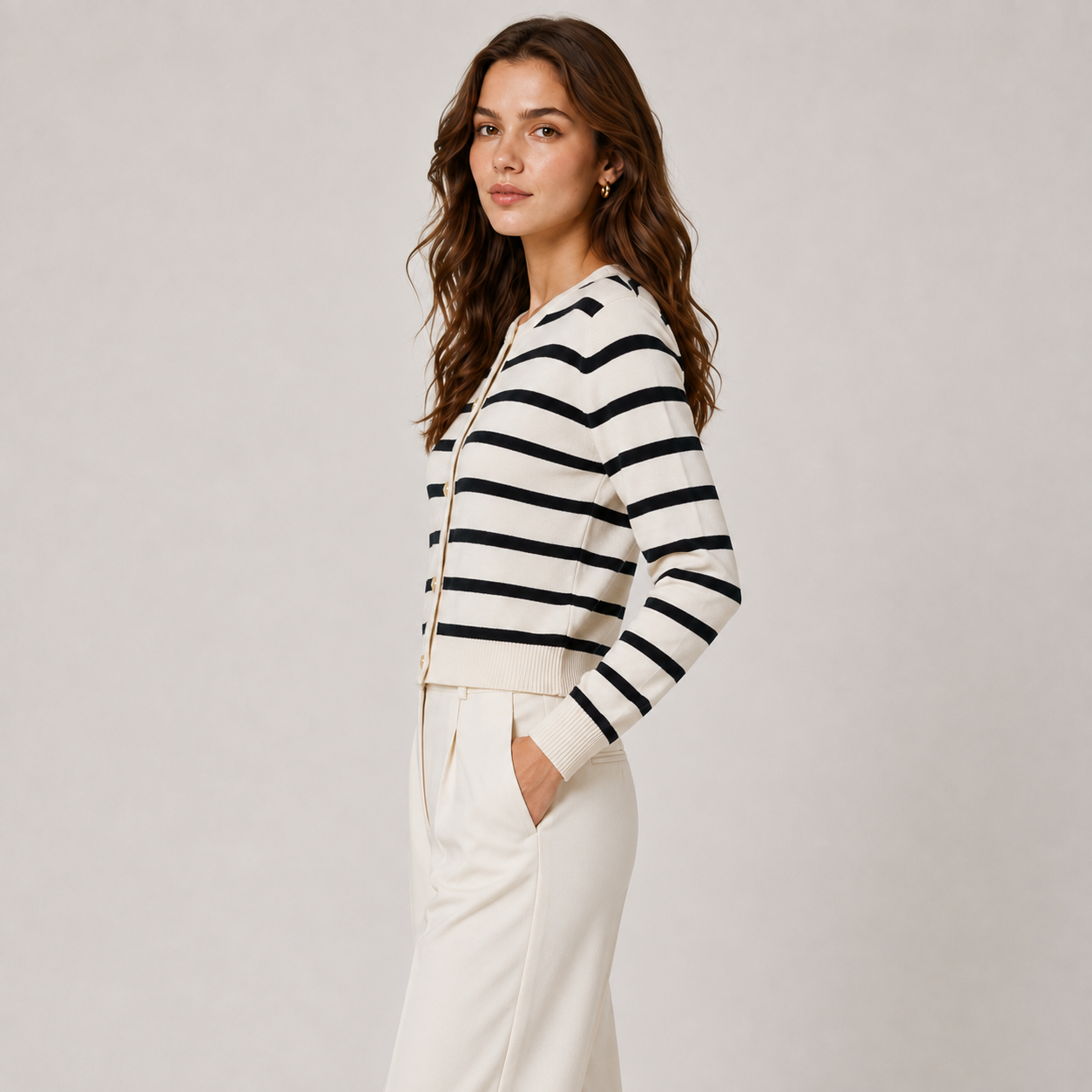 A woman modeling a cropped, long-sleeve knit cardigan with horizontal black and off-white stripes, paired with off-white trousers, against a neutral light gray studio background.