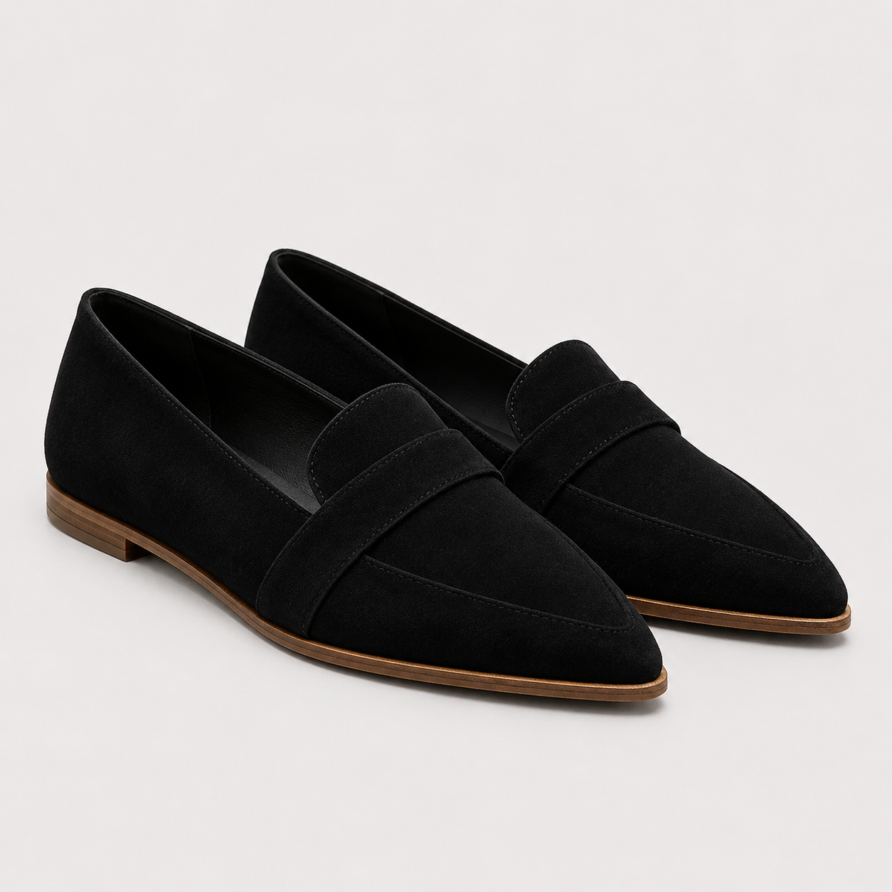 A pair of black suede penny loafers with pointed toes and low brown block heels, photographed on a light gray studio background.