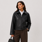 A woman models a black faux-leather zip-up jacket over a white collared shirt and brown trousers, holding a black handbag against a light gray studio background.