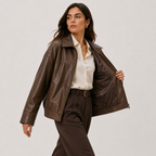 A woman models a brown faux-leather jacket, worn open over a cream button-down shirt and brown trousers, against a plain light gray studio background.