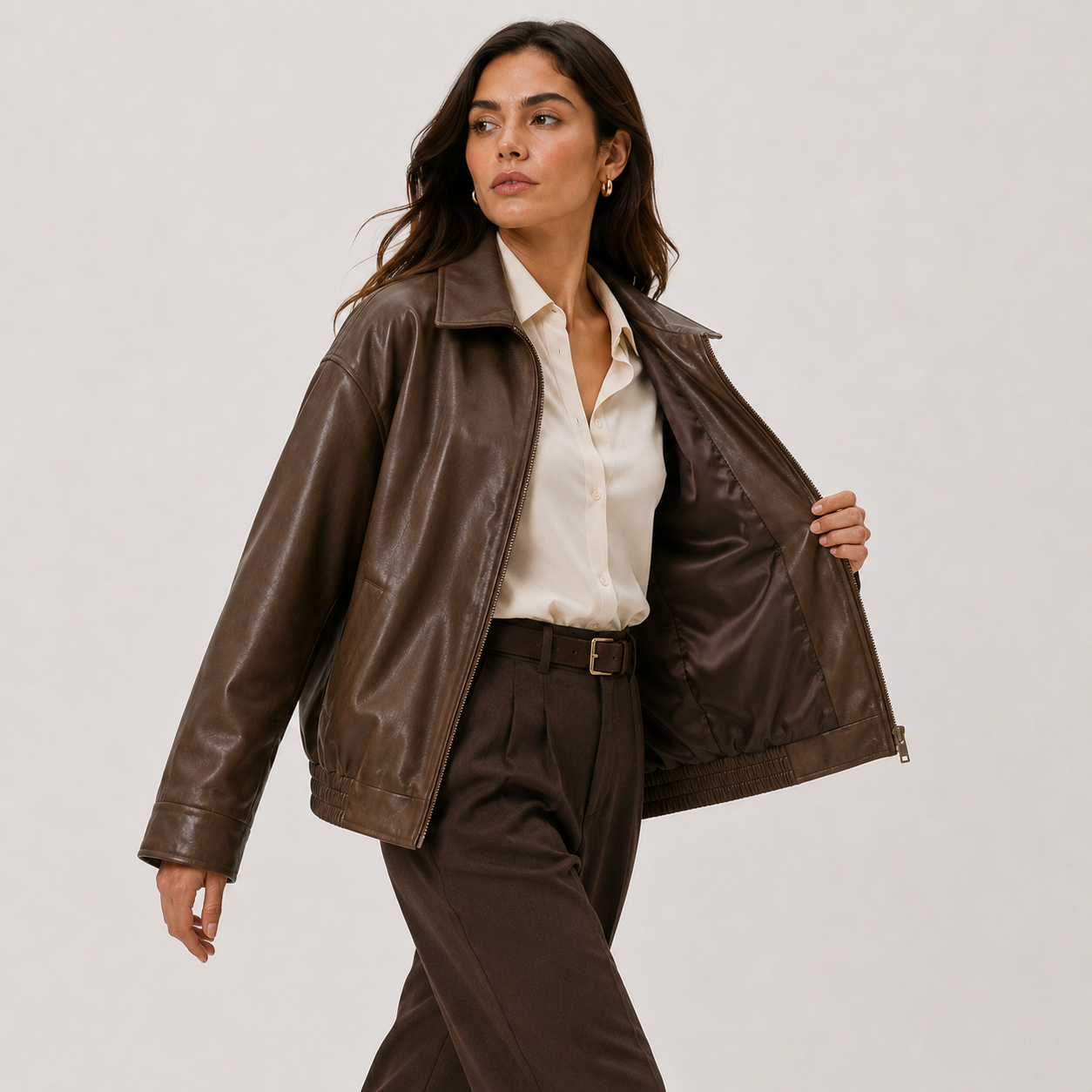 A woman models a brown faux-leather jacket, worn open over a cream button-down shirt and brown trousers, against a plain light gray studio background.