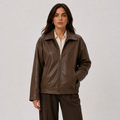 A woman modeling a dark brown faux-leather zip-up jacket over a cream collared shirt and dark brown trousers, photographed against a neutral light gray studio background.
