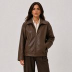 A woman modeling a dark brown faux-leather zip-up jacket over a cream collared shirt and dark brown trousers, photographed against a neutral light gray studio background.
