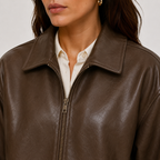 A close-up of a woman wearing a brown faux-leather zip-up jacket over a white collared shirt, cropped from the lower face to the chest against a light neutral background.