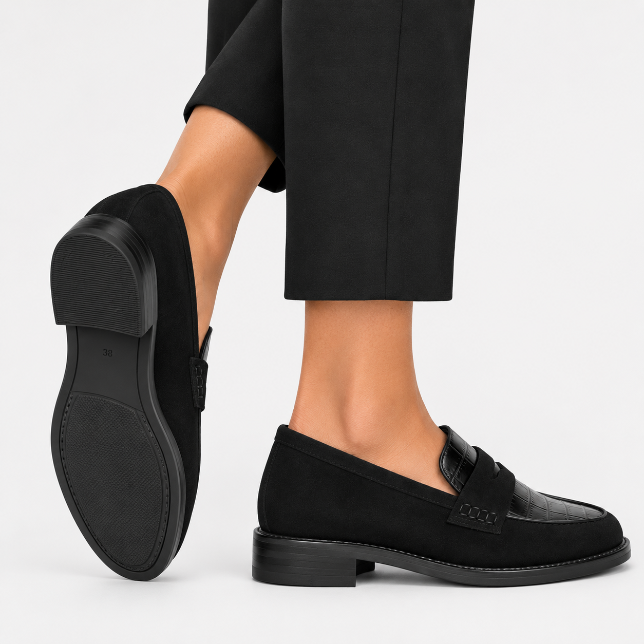 A pair of black suede and leather penny loafers shown on a model's feet, with one shoe turned to display the sole and the other shown from the side profile against a light gray background.