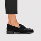 A black loafer shoe with a suede upper, crocodile-embossed toe cap, and low block heel, shown on a model wearing black trousers against a light gray background.