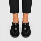 A pair of black crocodile-embossed leather penny loafers worn with black trousers, shown from a front-facing angle on a light gray background.