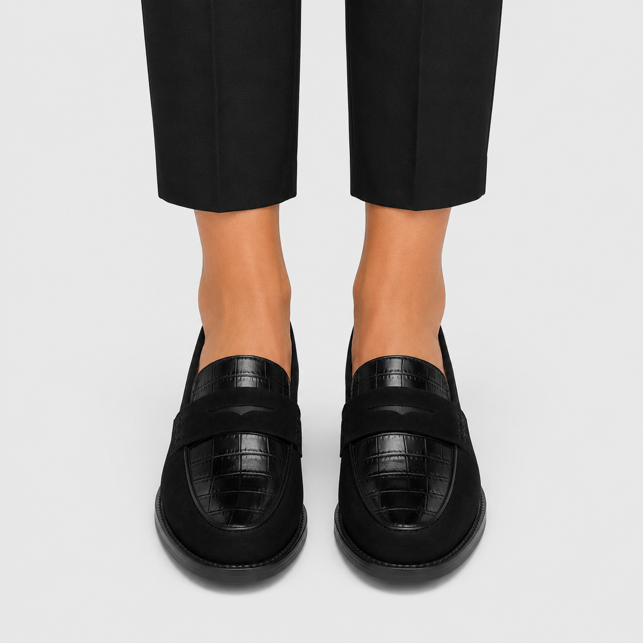 A pair of black crocodile-embossed leather penny loafers worn with black trousers, shown from a front-facing angle on a light gray background.