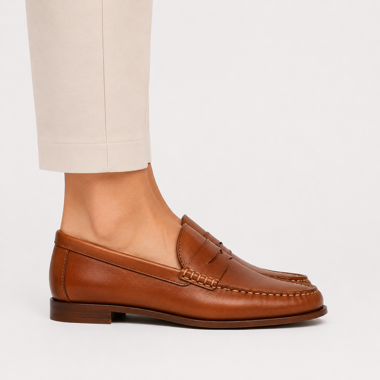 A person wearing a tan leather penny loafer with light beige trousers, shown from the side against a clean white background.