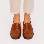 A pair of brown leather penny loafers worn with beige cropped trousers, shown from the front on a light gray studio background.