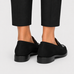Black slip-on loafers shown from the back on a model wearing black trousers, against a light gray studio background.