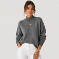 A woman wearing a gray ribbed mock neck sweater and white pants, standing against a light gray studio background.