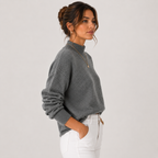 A woman wearing a gray knit mock neck sweater and white pants, shown in profile against a light gray studio background.
