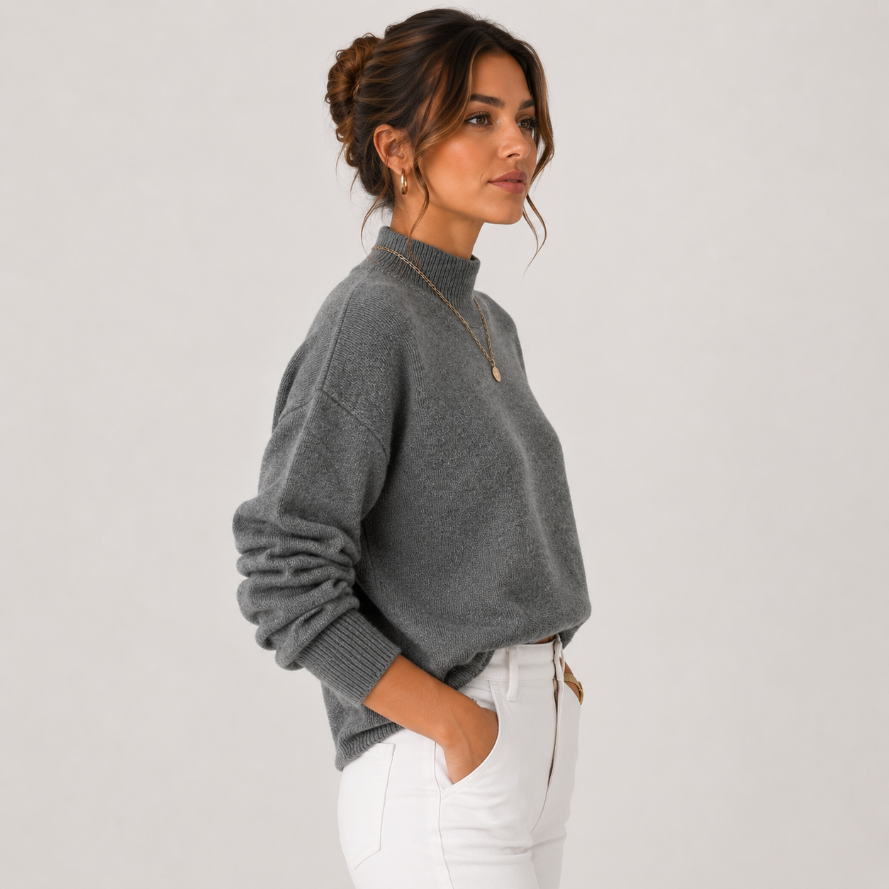 A woman wearing a gray knit mock neck sweater and white pants, shown in profile against a light gray studio background.