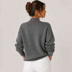 A woman shown from the back wearing a dark gray knit sweater with ruched sleeves and white pants, photographed against a plain light gray studio background.