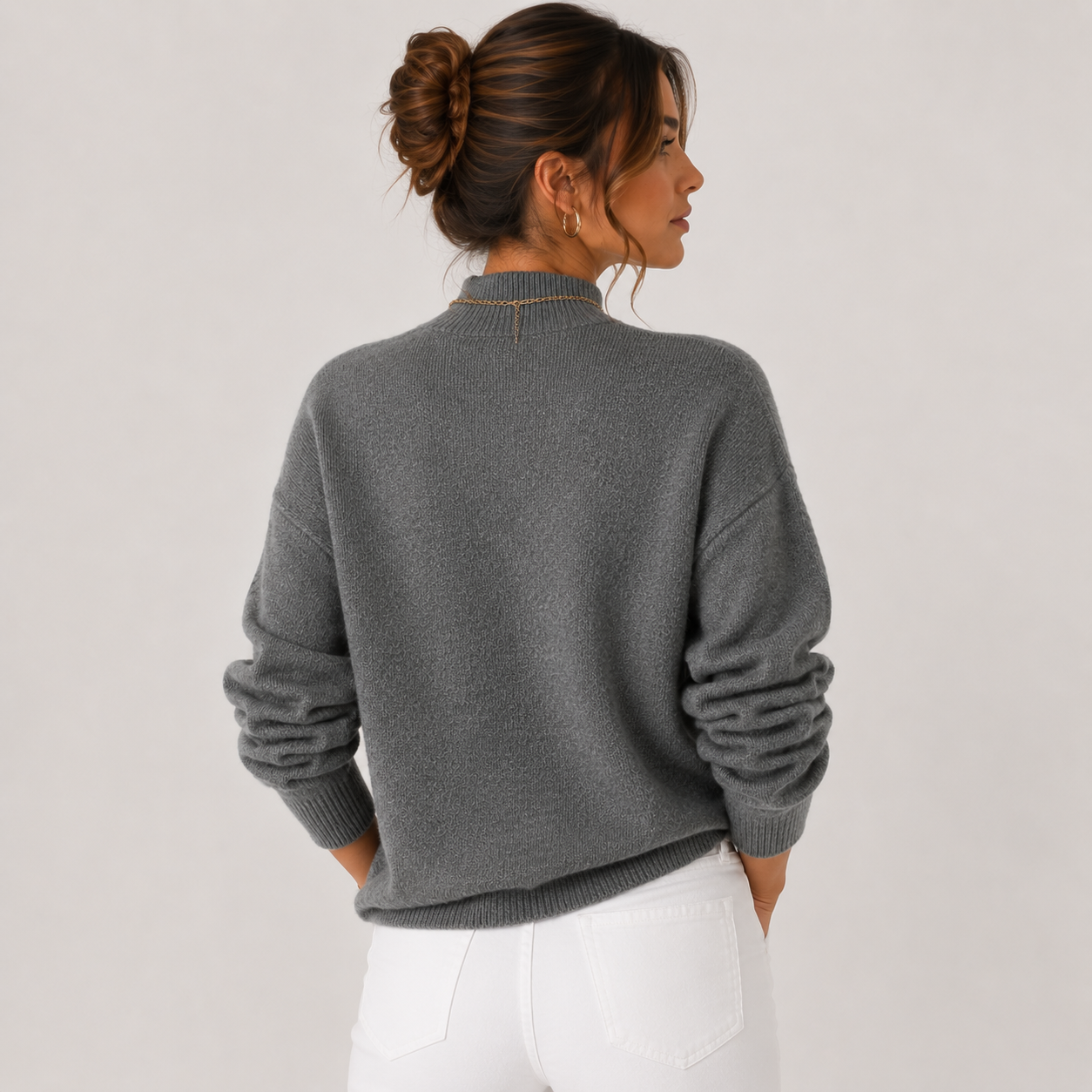 A woman shown from the back wearing a dark gray knit sweater with ruched sleeves and white pants, photographed against a plain light gray studio background.