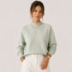 A woman wearing a light sage green mock neck knit sweater and white pants, accessorized with layered gold necklaces, rings, and a watch, against a solid pale pink background.
