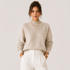A woman wearing a light beige knit mock neck sweater and white trousers, standing against a plain light gray studio background.