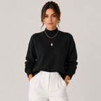 A woman wearing a black knit mock-neck sweater and white trousers, accessorized with layered gold necklaces and a gold watch, standing against a light gray studio background.
