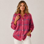A woman wearing a pink knit button-up cardigan with a blue and purple plaid pattern, paired with white pants, against a neutral light gray background.