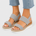 A pair of women's open-toe wedge sandals with textured light gray straps, shown on a model's feet with light blue jeans against a light gray studio background.