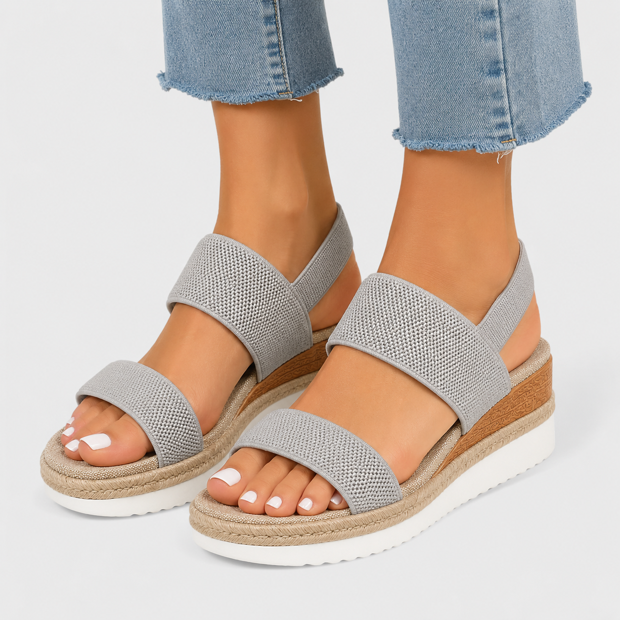 A pair of women's open-toe wedge sandals with textured light gray straps, shown on a model's feet with light blue jeans against a light gray studio background.