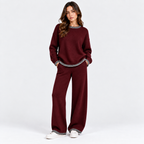 A woman modeling a matching burgundy knit lounge set consisting of a crewneck sweatshirt and wide-leg pants, both featuring a contrasting black and white patterned trim at the collar, cuffs, and hem, against a plain white studio background.