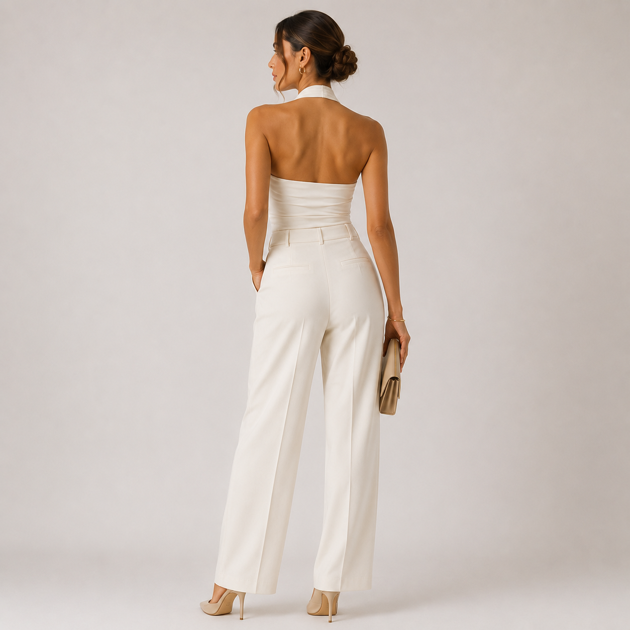 A woman is shown from behind wearing a white strapless halter-neck jumpsuit with wide legs, paired with beige heels and holding a small beige clutch, set against a plain light gray studio background.