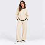 A woman modeling a cream-colored knit lounge set consisting of a crewneck sweatshirt and wide-leg pants, both featuring black beaded trim at the collar, hem, and cuffs, against a plain white studio background.
