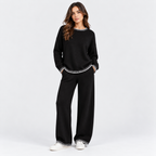 A woman modeling a black knit lounge set with white trim details, consisting of a crewneck sweatshirt and matching wide-leg pants, paired with white sneakers on a light gray studio background.