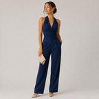 A woman modeling a navy blue halter-neck jumpsuit with a cinched waist and wide legs, holding a beige clutch against a light gray studio background.