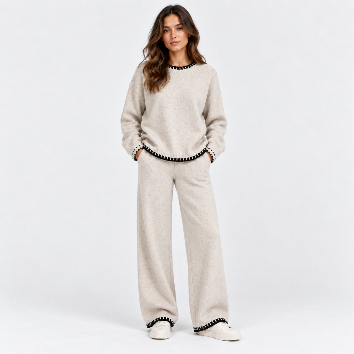 A woman modeling a matching beige knit lounge set with black beaded trim along the neckline, cuffs, and hem, paired with white slip-on shoes, against a clean white studio background.
