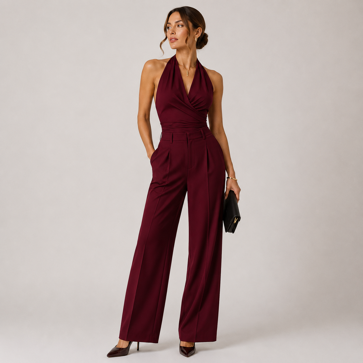 A woman models a sleeveless halter-neck jumpsuit in deep burgundy with a crossover V-neck bodice, defined waist, and wide-leg trousers, paired with dark brown heels and a small black clutch, set against a plain light gray studio background.