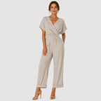 A woman modeling a light beige wrap-style jumpsuit with short sleeves, a twisted waist detail, and wide cropped legs, paired with nude high-heeled sandals against a light gray studio background.