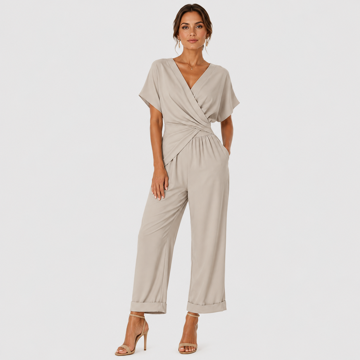 A woman modeling a light beige wrap-style jumpsuit with short sleeves, a twisted waist detail, and wide cropped legs, paired with nude high-heeled sandals against a light gray studio background.