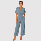 A woman modeling a dusty blue wrap-style jumpsuit with short flutter sleeves, a twisted waist detail, and wide cropped legs with rolled cuffs, photographed against a light gray studio background.