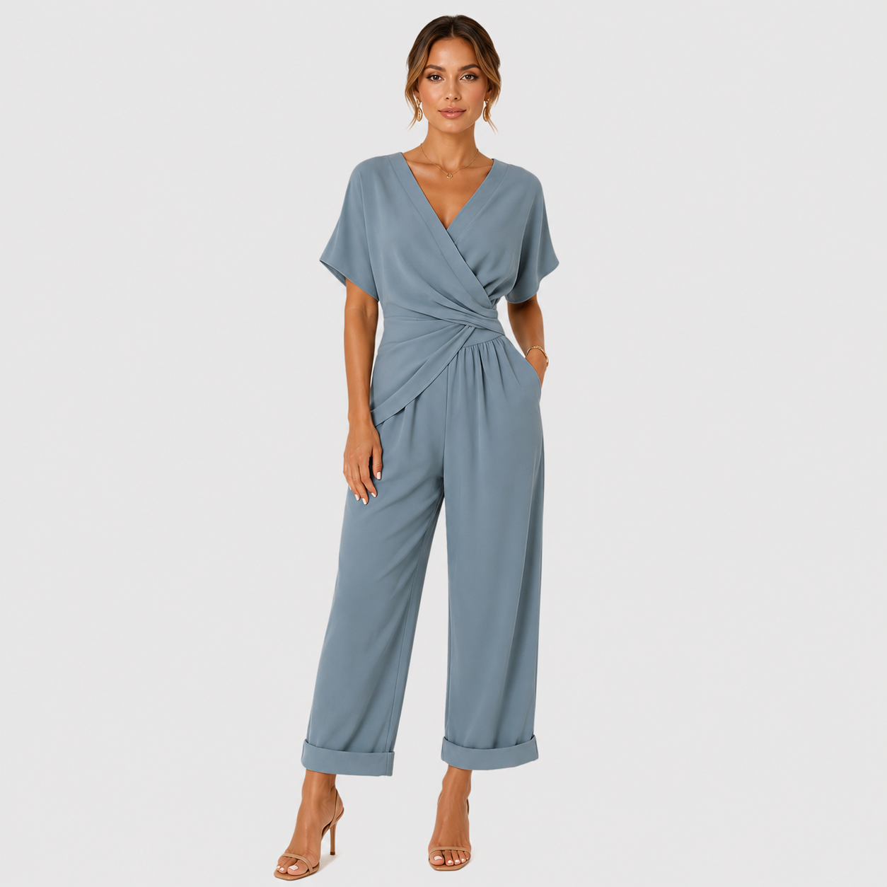 A woman modeling a dusty blue wrap-style jumpsuit with short flutter sleeves, a twisted waist detail, and wide cropped legs with rolled cuffs, photographed against a light gray studio background.