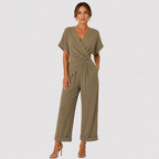 A woman modeling an olive green wrap-style jumpsuit with short sleeves and wide cropped legs, standing against a light gray studio background.