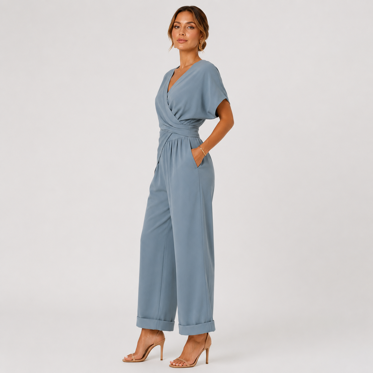 A woman modeling a dusty blue wrap-style jumpsuit with short sleeves, side pockets, and wide cropped legs, standing against a plain light gray studio background.